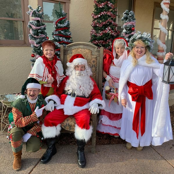 Santa Claus, Mrs. Claus, an elf, and two women in festive attire pose outdoors with Christmas trees.