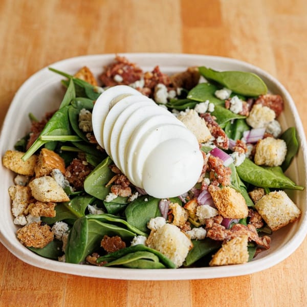Spinach salad with sliced hard-boiled eggs, croutons, bacon bits, and feta cheese in a white bowl.