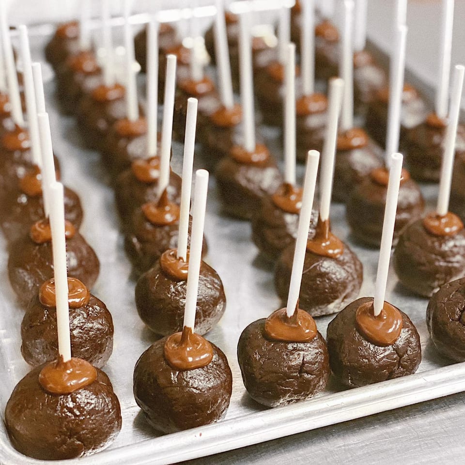Rows of chocolate cake pops with caramel drizzle on a silver tray at eats & sweets bakery.