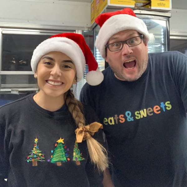 Two eats & sweets employees in Santa hats, one with a Christmas tree shirt, smiling at the camera.